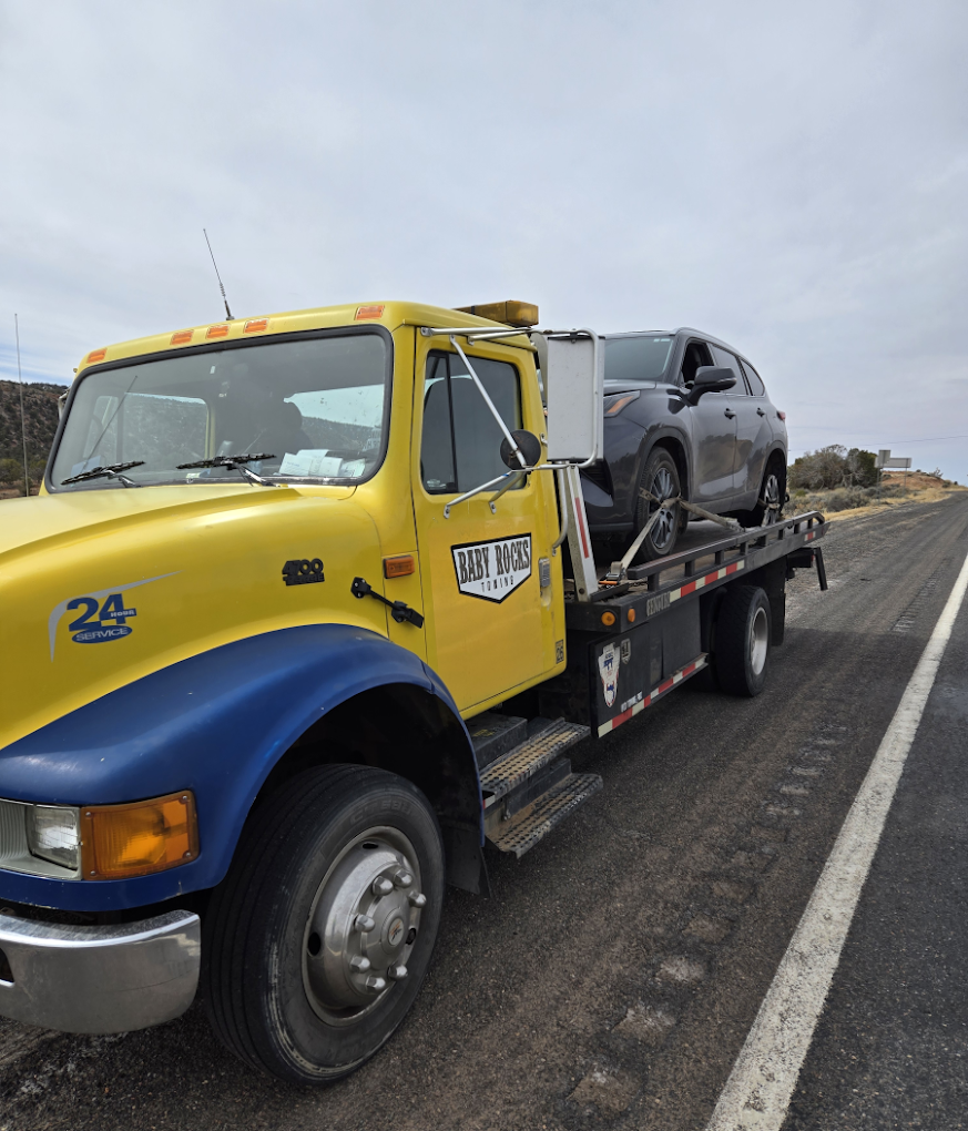 A yellow Baby Rocks Towing flatbed truck providing 24-hour reliable Towing services in Kayenta, AZ like roadside vehicle transport and recovery services on a highway.